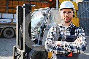 forklift driver in protective vest and forklift standing at warehouse of freight forwarding company, smiling