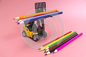 A forklift truck holds colored pencils on a platform and loads them onto a stack from below. The concept of logistics and delivery