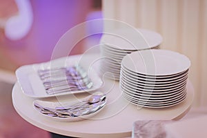 Forks and knives and stack of white dishes in the background. Stack of white plates