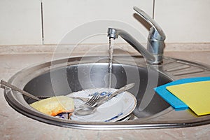 Forks, spoons, plates of different sizes, rags and a sponge in the sink under the tap