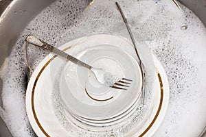 Forks, spoons, plates of different sizes in the sink under running water.