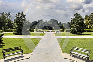 Formal garden with straight paths and benches and a fountain and beautiful trees under cloudy sky with rainbow