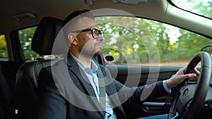 Formally dressed bearded man in glasses driving a car in sunny weather