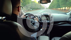 Formally dressed bearded man in glasses driving a car in sunny weather. View from the back seat of a car