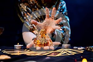 The fortune teller holds a glass pyramid with gold in her hands and casts a spell over it. There are Tarot cards on the table.