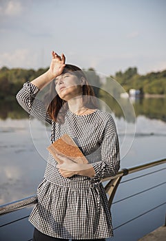 A forty-year-old woman in a plaid tunic stands with a book in her hands against the river