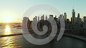 Forwards fly above river. Aerial view of Brooklyn bridge against bright sunset sky. Skyscrapers in Financial District