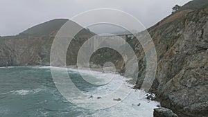 Forwards fly along rugged rocky coast. Concrete arch road bridge over valley at sea coast. California, USA