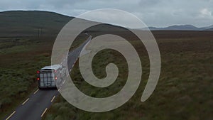 Forwards fly behind vehicle with horse trailer at dusk. Narrow road in countryside surrounded by pastures and grasslands