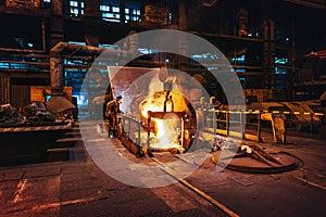 Foundry workshop interior, molten iron pouring from blast furnace into ladle container and workers founders control