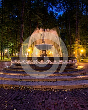 A fountain with blurred streams of water in a night park illuminated by lanterns with a stone pavement, trees and benches