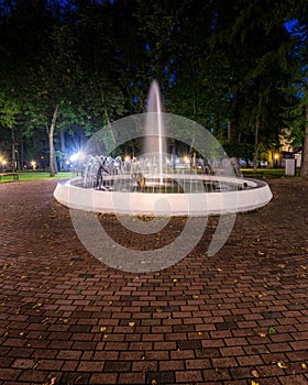 A fountain with blurred streams of water in a night park illuminated by lanterns with a stone pavement, trees and benches