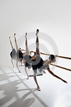 Four ballet dancers line up at barre, performing identical pose with outstretched leg, training against grey studio