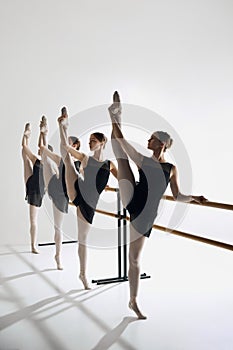 Four ballet dancers line up at barre, performing identical pose with outstretched leg, training against grey studio