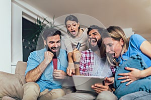 Four casual happy friends laughing shopping online together in a tablet sitting on a sofa in the living room