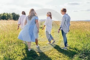 Four cheerful smiling women walking by the high green grass meadow hill, laughing and chatting with each other during a sunset