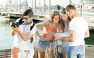Four cheerful traveling people searching for direction using paper map on waterfront in town