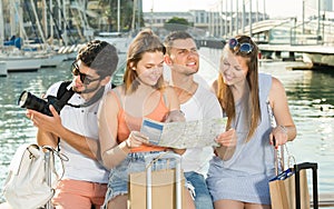 Four cheerful traveling young people searching for direction using paper map on waterfront in town