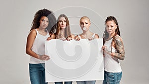 Four diverse women looking at camera while holding, standing with blank banner in their hands isolated over grey