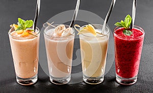 Four drink glass with smoothies, currant, raspberries, peach, banana, apple on black stone background. Summer cold drink and