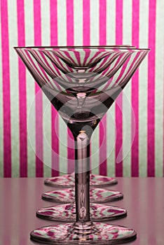 Four empty glasses for martini and vermouth standing in line with white and pink striped background.