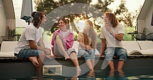 Four friends sit near the sunbeds by the pool and have fun chatting with each other against the background of the sunset