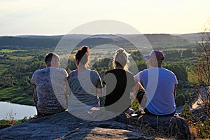 Four friends sitting on edge of mountain at sunset