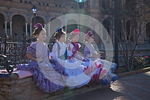 Four girls dancing flamenco, looking distracted, in typical flamenco costumes sitting on a bench in a nice square in Seville,