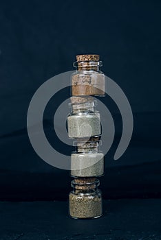 Four glass bottles with green, beige and brown powder inside placed one on one vertically on black background. Spices, teas, food