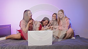 Four happy pretty young women in pajamas sits on bed, eats popcorn and watch a movie at bachelorette party