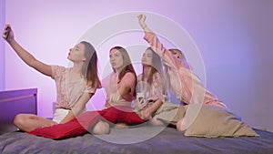 Four happy pretty young women in pajamas sits on bed, sings and records video on their phones at bachelorette party