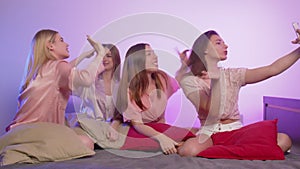 Four happy pretty young women in pajamas sits on bed, sings and records video on their phones at bachelorette party