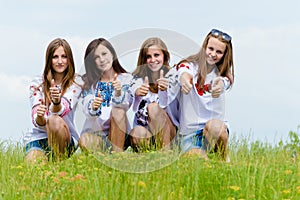 Four happy young women friends showing thumbs up in green grass over blue sky