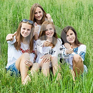 Four happy young women friends smiling & showing thumbs up in green grass