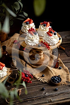 Four homemade white mini desserts pavlova on wooden cake stand with whipped cream and assorted berries on rustic table