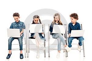 Four kids in denim clothes sitting on chairs and using laptops on white.