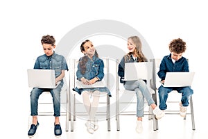 Four kids in denim clothes sitting on chairs and using laptops on white.