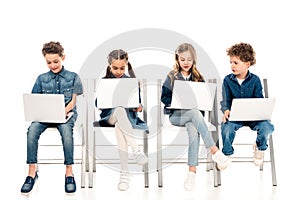 Four kids in denim clothes sitting on chairs and using laptops on white. photo