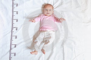 A four month happy baby in pink white clothes lying on a bed on which a measuring ruler for growth is drawn