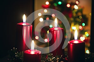 Four red candles burning on advent wreath with blurred colorful christmas tree with glowing lights in the background on evening