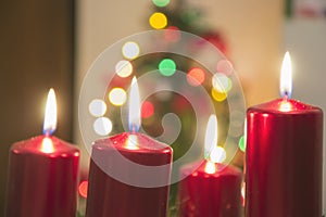 Four red candles burning on advent wreath with blurred colorful christmas tree with glowing lights in the background on evening