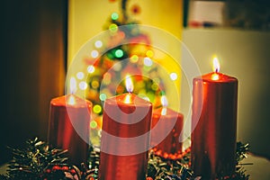Four red candles burning on advent wreath with blurred colorful christmas tree with glowing lights in the background on evening