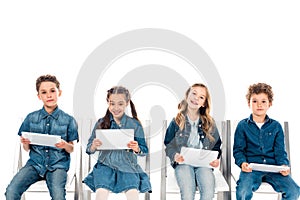 Four smiling kids in denim clothes sitting on chairs and using digital tablets isolated on white.