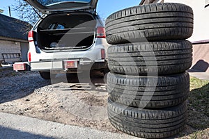 Four summer tyres lying on land next to opened trunk of silver suv car, seasonal tire change