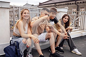 Four teenagers are sitting on the parapet on the roof of the house and eating Asian food. Two young couples snack on fast food and photo