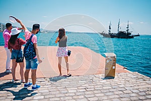 Four tourists waving at an old black pirate boat which is passing by next to a modern marine port