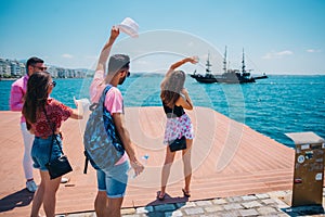 Four tourists waving at an old black pirate boat which is passing by next to a modern marine port