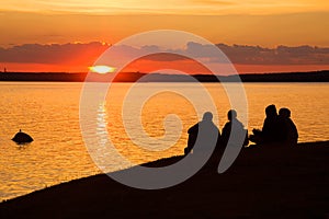 Four unknown human silhouettes are sitting on the shore and watching a beautiful sunset