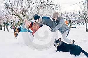 Four warm young Caucasian girls and a dog with one eye of each color, play in the snow making big snowballs