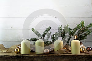 Four white candles, one of them burning on the first advent christmas decoration with spruce twigs, baubles and a golden ribbon o